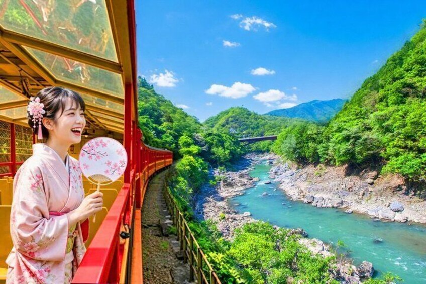 A person in traditional attire holding a fan, overlooking a river with a bridge and lush greenery. 1