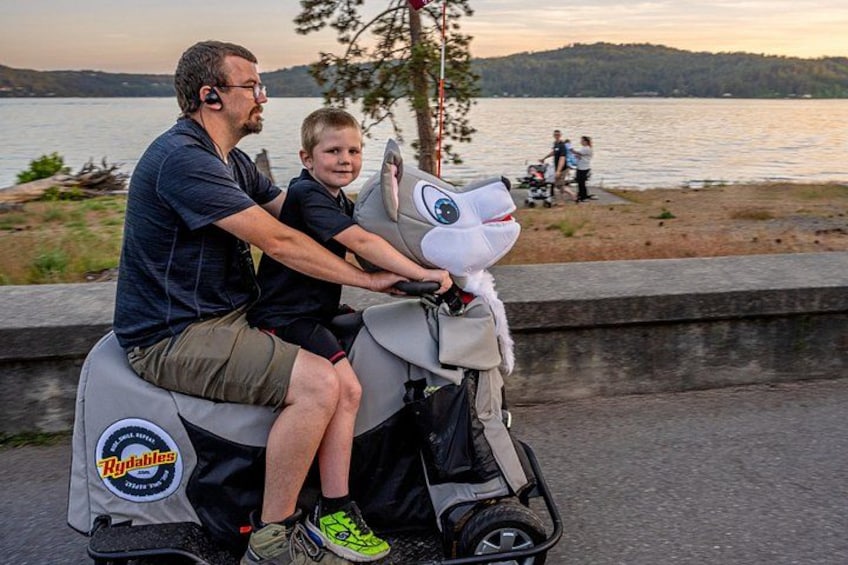 A man and a child on a scooter with a large animal head decoration, near a body of water with people in the background. 3