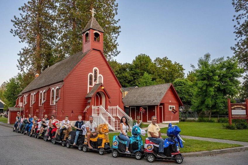 A red church with a steeple, a small building next to it, and a group of people on colorful scooters. 4