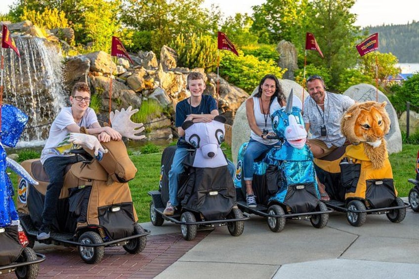 People riding animal-shaped carts in a park with a waterfall and flags. 2