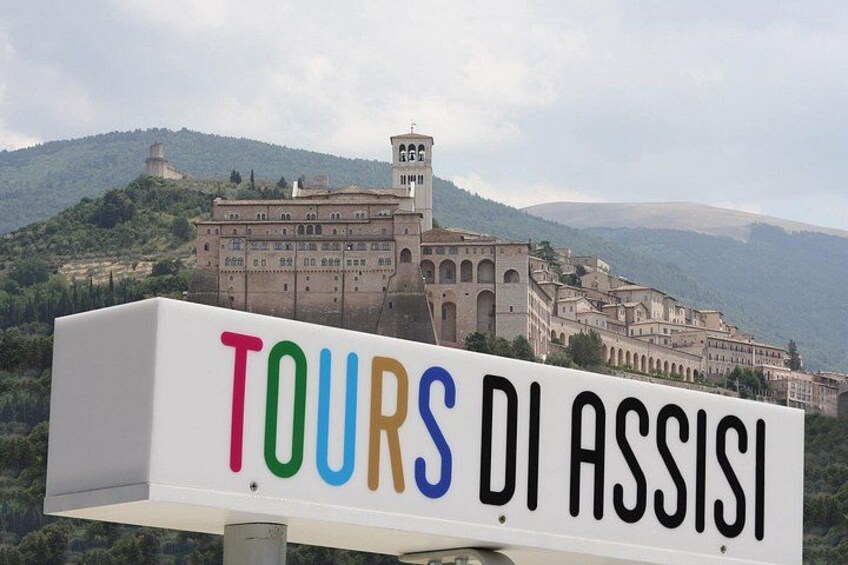 A sign with 'TOURS DI ASSISI' in colorful letters, a historic building, and a mountainous landscape in the background. 1