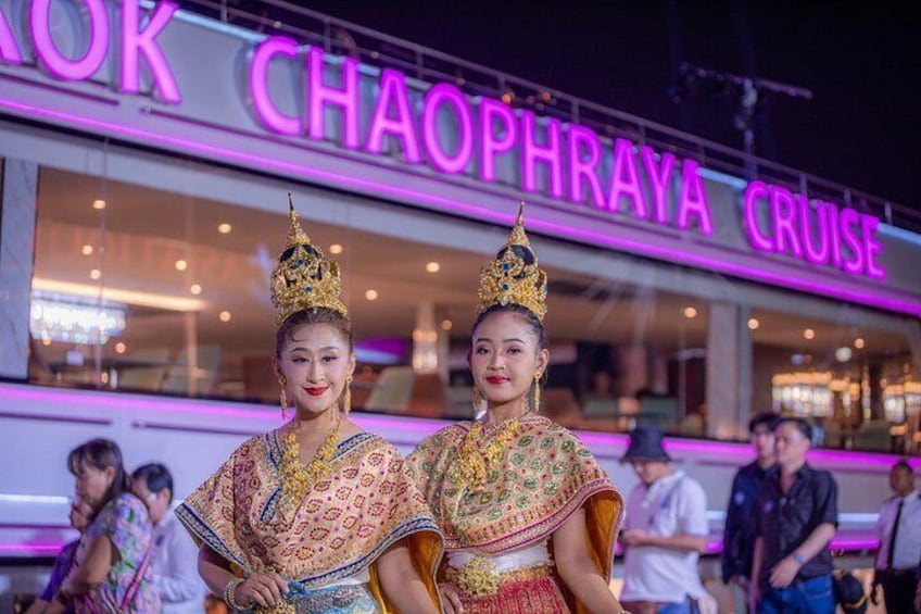 Two individuals in traditional attire with elaborate headdresses in front of a brightly lit building with signage. 3