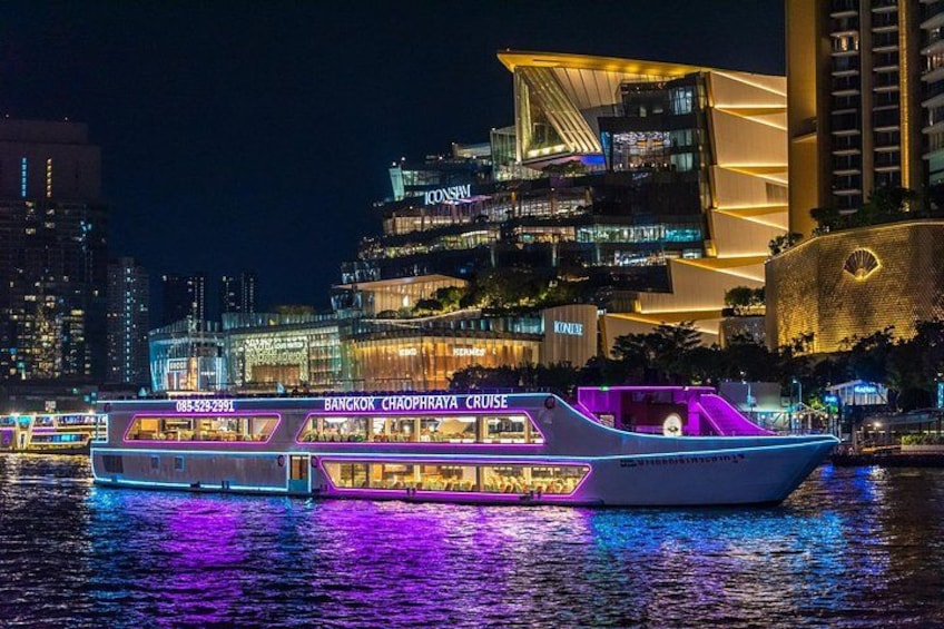 Ein hell beleuchtetes Kreuzfahrtschiff mit Neonlichtern auf einem Fluss bei Nacht, im Hintergrund moderne Gebäude. 1