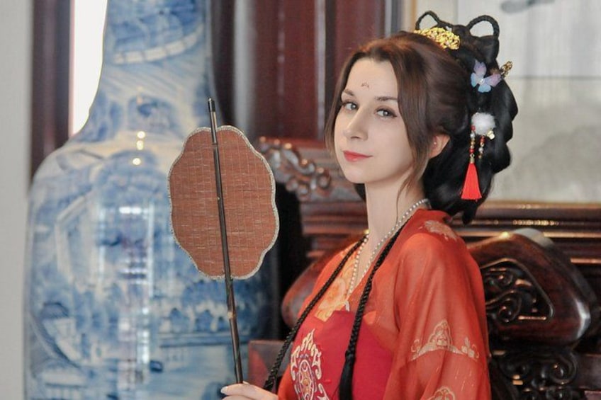 A person in traditional attire holding a fan, standing in front of a large blue vase. 2