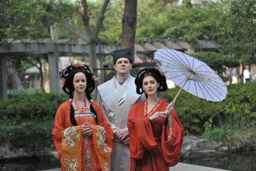 Three people in traditional attire, one holding an umbrella, standing in a garden with trees and a stone bridge in the background. 4