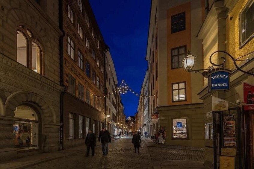 Eine gepflasterte Straße gesäumt von historischen Gebäuden, eine Straßenlaterne und ein Schild für ein Geschäft namens 'TODAKS HANDEL'. 3