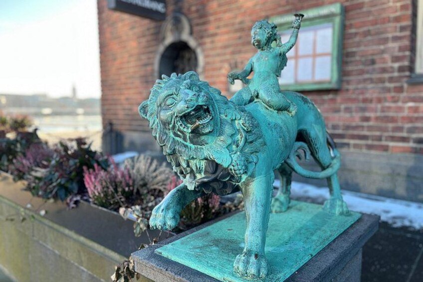 A bronze statue of a lion with a rider on a pedestal, set against a brick building with a window and a sign. 2