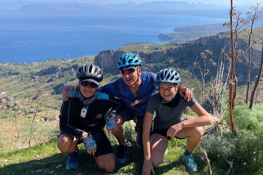 Tre personer med hjälmar och cykelkläder poserar på en gräsklädd kulle med en vacker utsikt över en kustlinje och berg i bakgrunden. 1