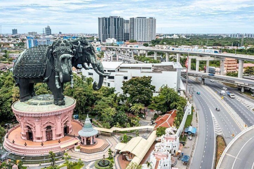 A large elephant statue on a pink pedestal, surrounded by a park with a fountain, in an urban area with roads and buildings. 3