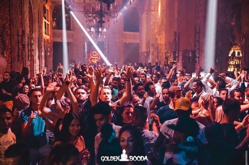 Evento interior concurrido donde personas levantan las manos, un candelabro y un letrero con el texto 'GOLDEN BOODA'. 4