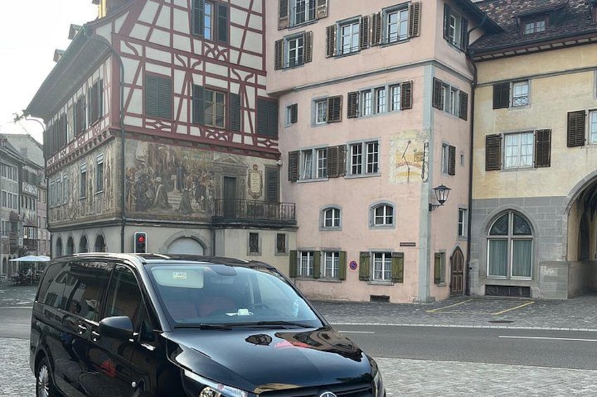 A black car parked on a cobblestone street, historic buildings with murals and traditional architecture in the background. 5
