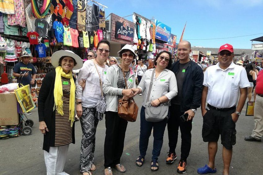 A group of people standing in a busy outdoor market with various shops and colorful items in the background. 2