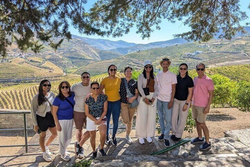 Eine Gruppe von Personen steht auf einem Steinweg, im Hintergrund ein Weinberg und Hügel. 1