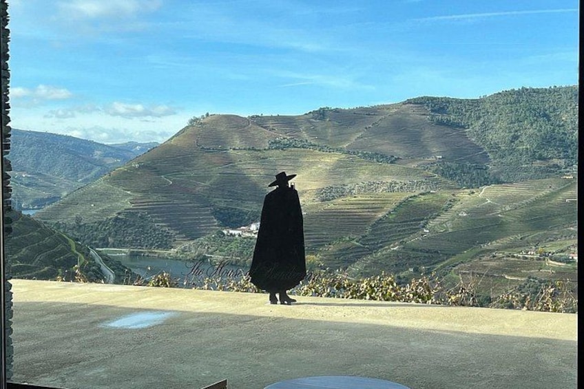 A person in a long black coat and hat standing on a road with a view of terraced hills and a bridge in the distance. 5