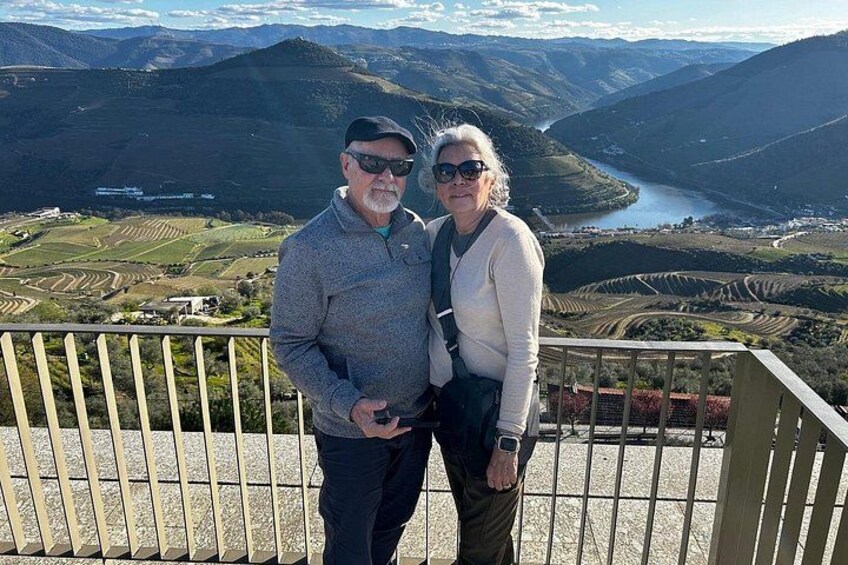 Two people standing on a balcony overlooking a valley with terraced fields and a river. 2