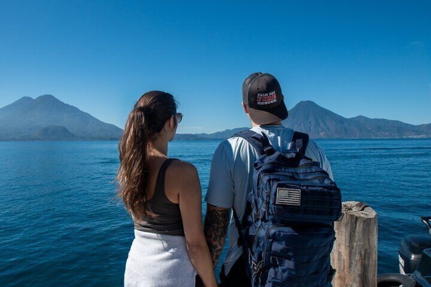 A couple standing on a dock overlooking a calm body of water with mountains in the background. 1