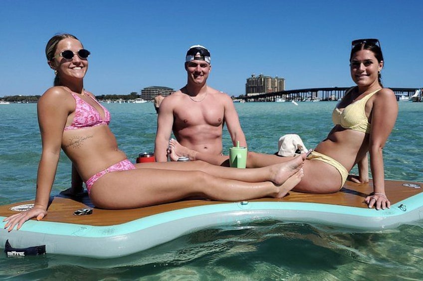 Drie personen op een drijvend platform in helder water, met een steiger op de achtergrond. 2