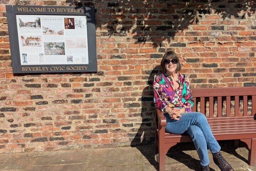 A woman sitting on a bench in front of a brick wall with a sign that reads "Welcome to Beverley" and "Beverley Civic Society". 1