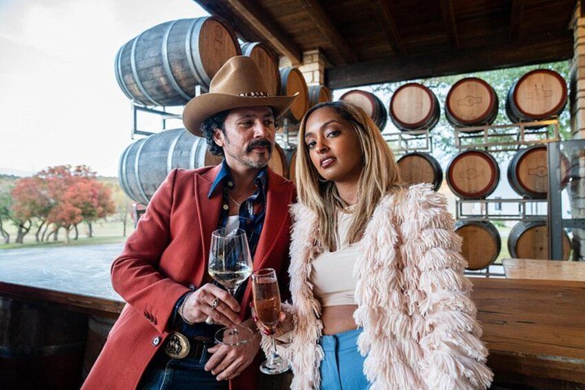 A man and a woman standing in a rustic setting with wooden barrels in the background. 2