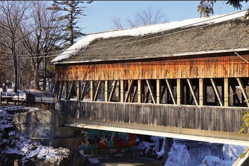 A wooden covered bridge with snow on the roof and a mural on the lower wall. 5