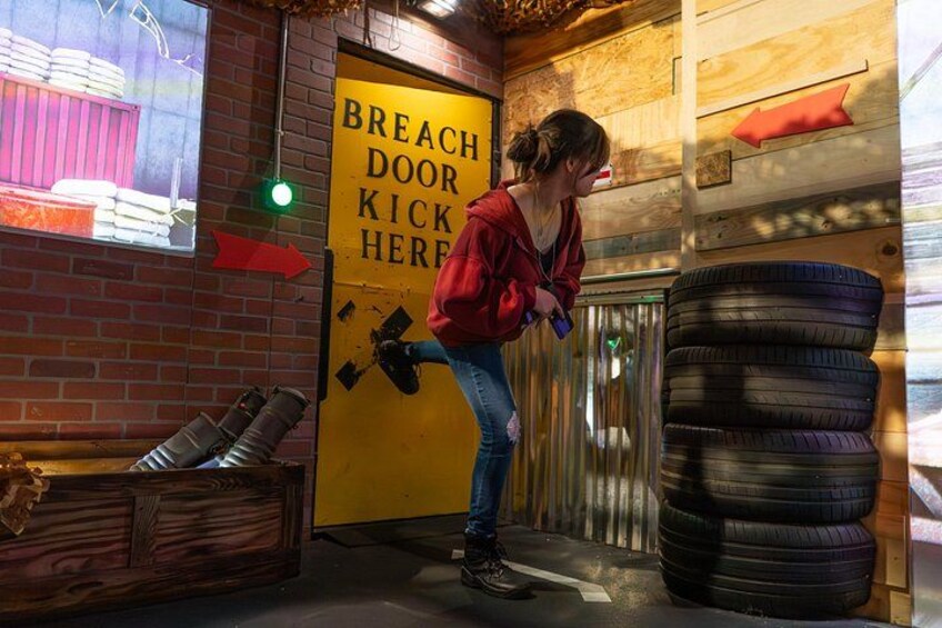 Una persona pateando un neumático en un área de juego de derribo de puertas, con un cartel que dice 'BREACH DOOR KICK HERE' y una pila de neumáticos. 4