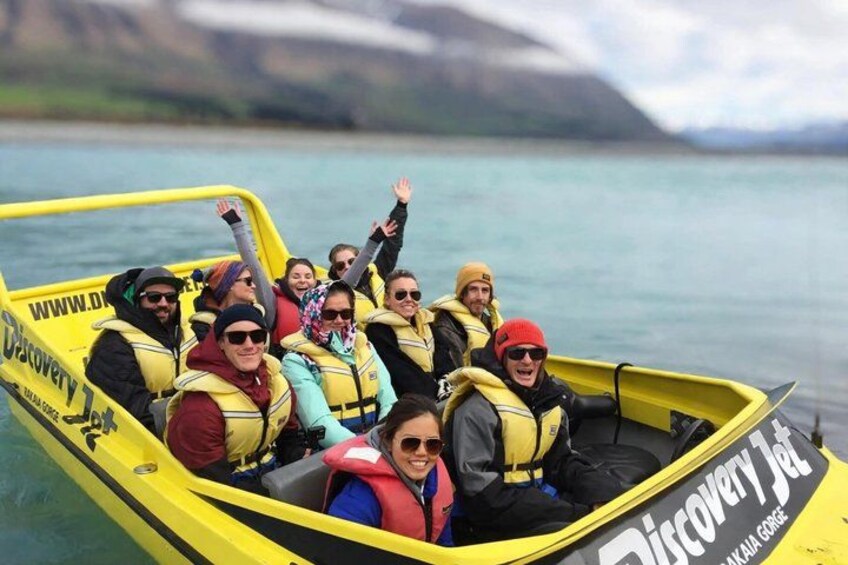 A group of people on a yellow boat with life jackets, on a calm body of water with mountains in the background. 4