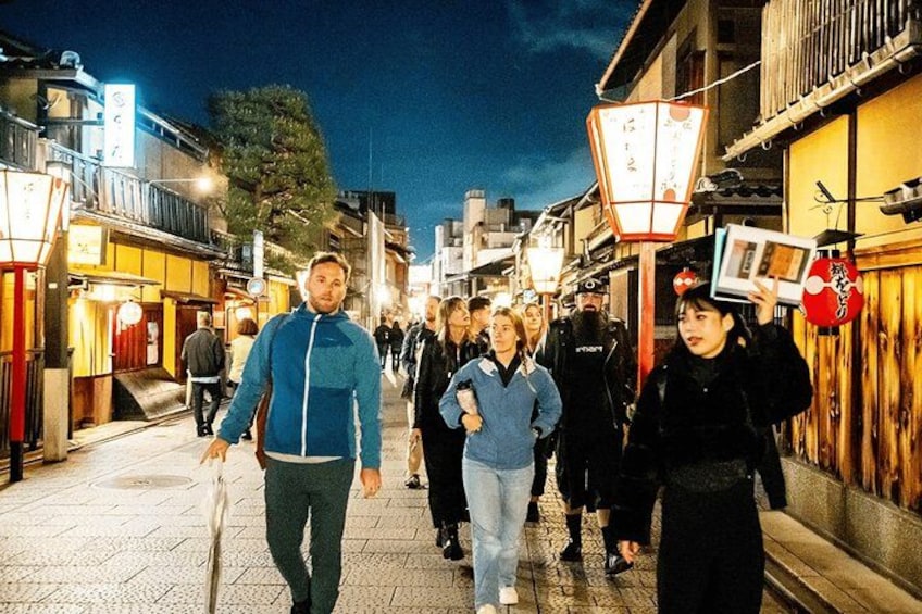 A nighttime street scene with people walking, illuminated by street lamps and shop signs. 2