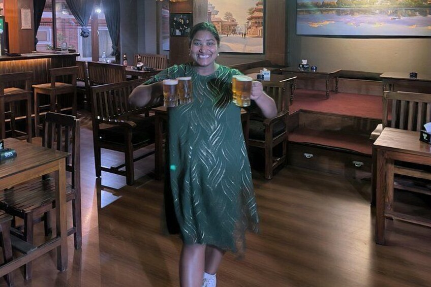 Een persoon in een groene jurk die een biertje vasthoudt in een restaurant met houten meubels en een groot scherm waarop een mooi landschap te zien is. 4