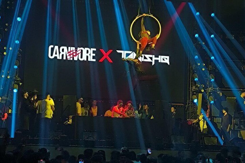 Een podium met een groot spandoek waarop 'Carnivore X Tansha' staat en optreders in gele outfits. 2