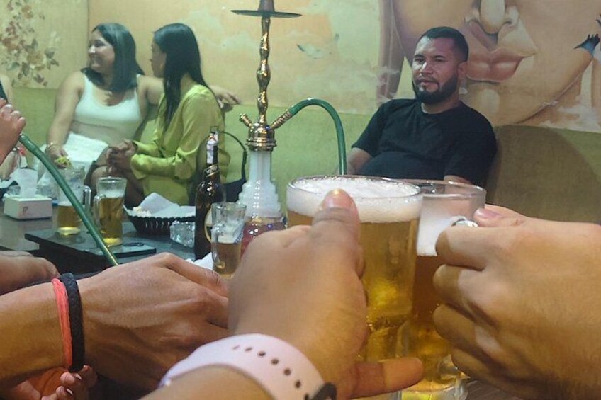 Un groupe de personnes trinquant avec des verres de bière, une pipe à eau et une bouteille posés sur une table. 4