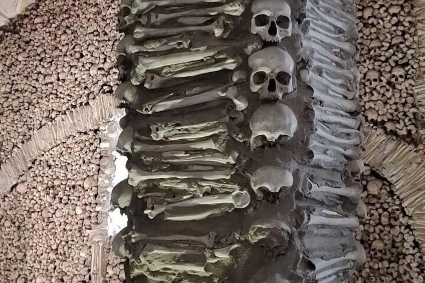 Una serie de cráneos y huesos humanos dispuestos en fila, con suelo rocoso y paredes de piedra. 3
