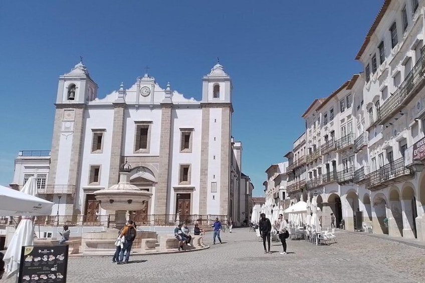 Una iglesia histórica con un campanario, una plaza adoquinada y gente caminando. 2