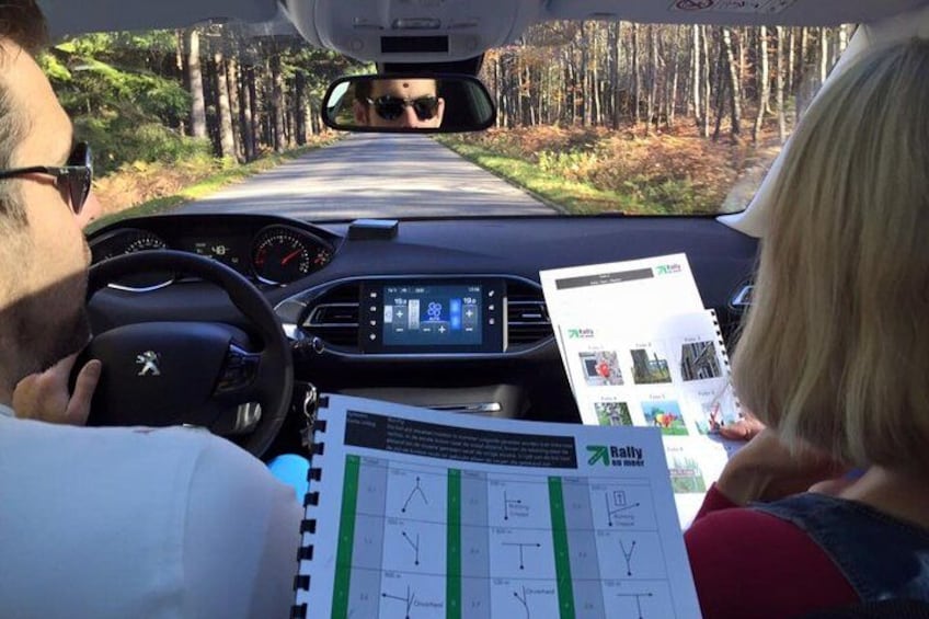 A car interior with a person driving, a passenger holding a Rally magazine, and a forested road ahead. 1