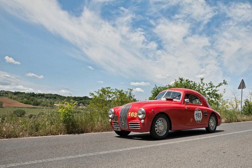 A red vintage car with the number 163 on its side, driving on a road with a rural landscape in the background. 2