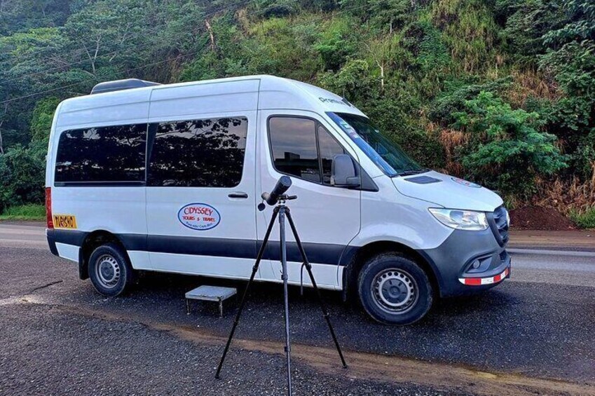 A white van with a tripod and a camera on the side, parked on a road with greenery in the background. 5
