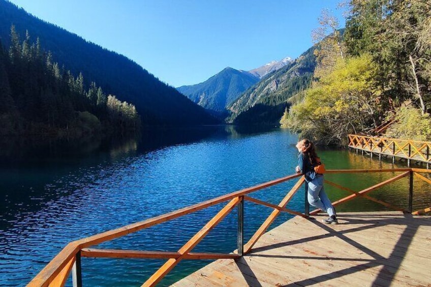 A person standing on a wooden dock with a railing, overlooking a calm lake surrounded by mountains. 1