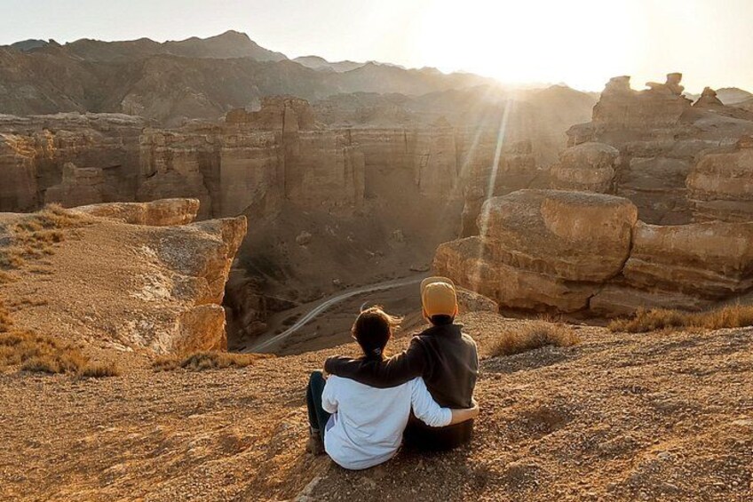 Two people sitting on a rocky outcrop overlooking a canyon with a winding road. 4