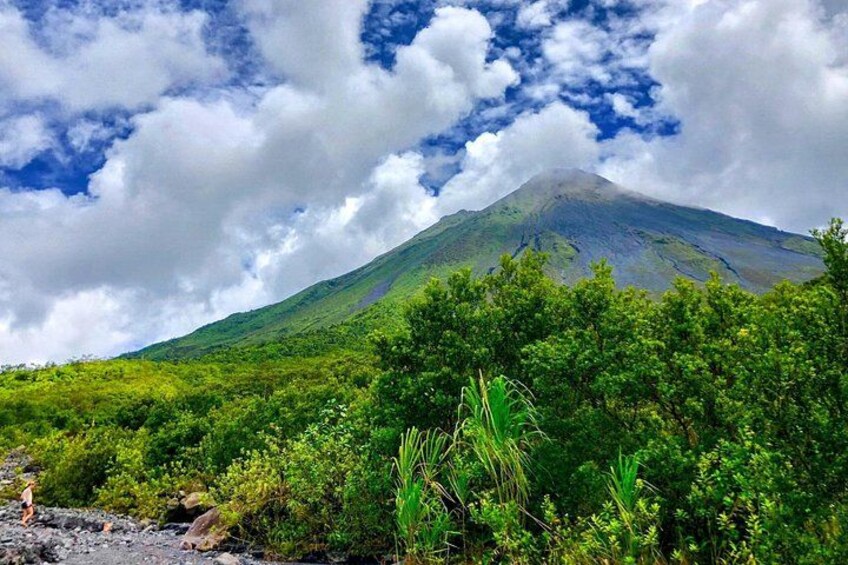A volcano with a cloud-covered peak, lush green vegetation in the foreground, and a partly cloudy sky. 3