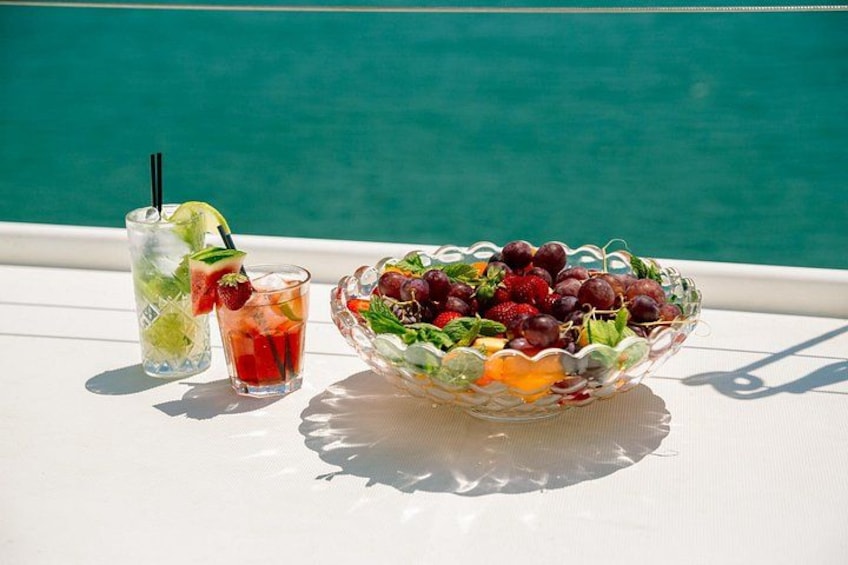 A glass of iced drink with a straw and a bowl of mixed fruit on a white table by a pool. 3