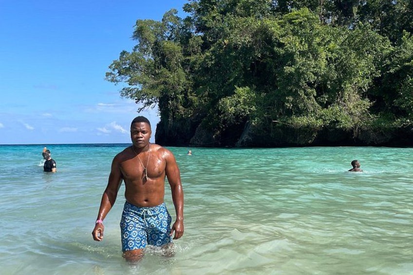A man in blue patterned swim trunks standing in clear turquoise water at a beach with lush green trees in the background. 4