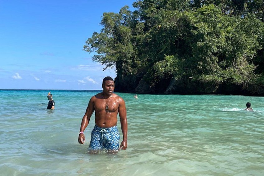 A man in blue patterned swim trunks standing in clear turquoise water at a beach with lush green trees in the background. 3
