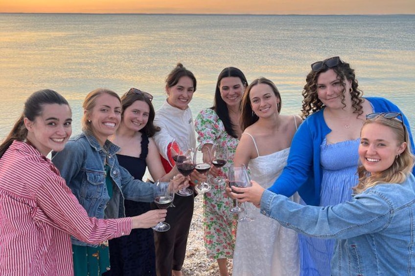 A group of people toasting with wine glasses by the sea during sunset. 3
