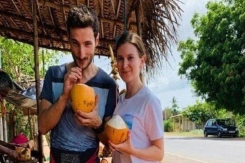 Two people holding coconuts, a thatched roof structure, and a parked car. 1