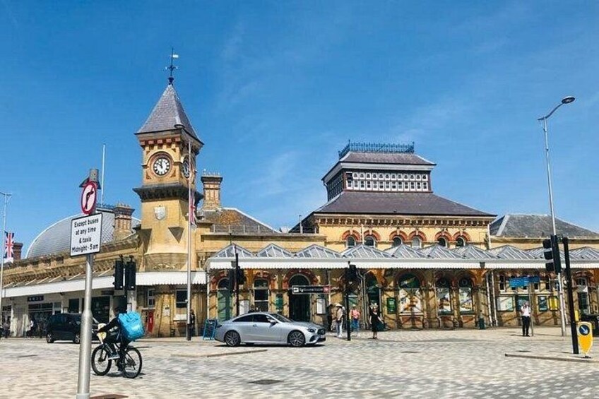 A historic train station with a clock tower, a covered waiting area, and a vintage car parked in front. 1