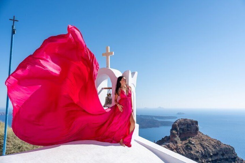 A woman in a red dress on a white platform with a cross and a bell in the background, overlooking a coastal landscape. 4