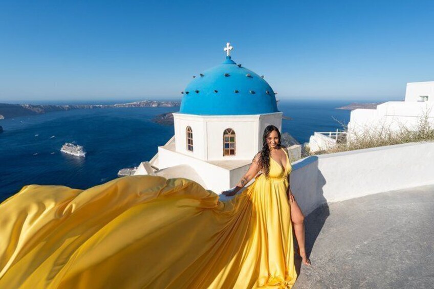 A woman in a yellow dress standing near a blue-domed building with a view of the sea. 1