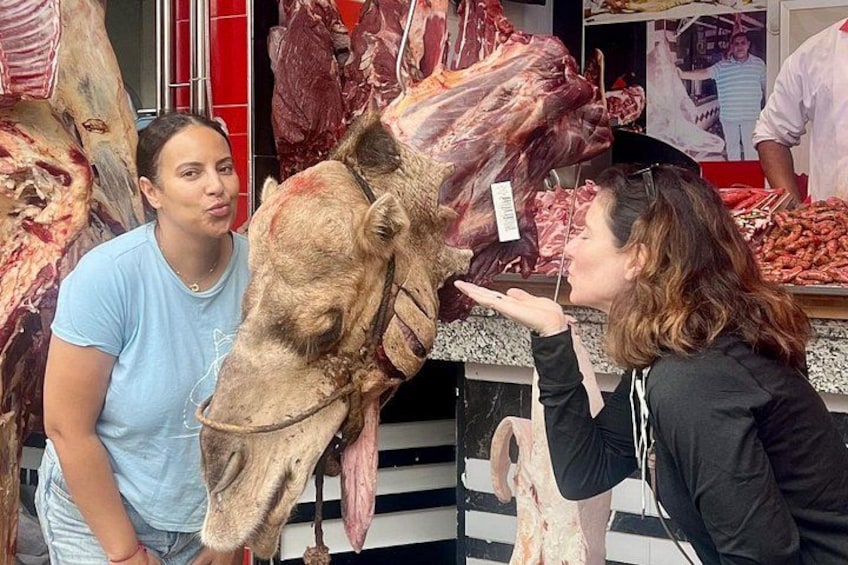 Une personne nourrit un chameau sur un marché, avec diverses viandes exposées en arrière-plan. 2
