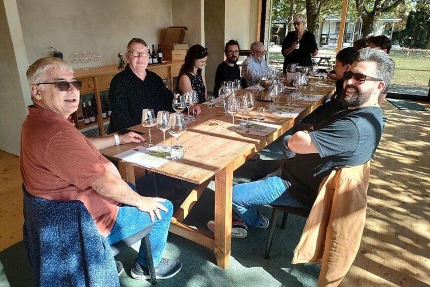 A group of people seated at a wooden table outdoors, with wine glasses and bottles visible. 3