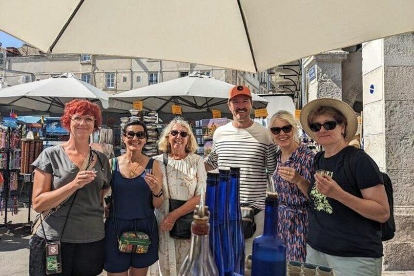 Un groupe de personnes se tient dans un marché en plein air, avec des parasols et des étals en arrière-plan. 5