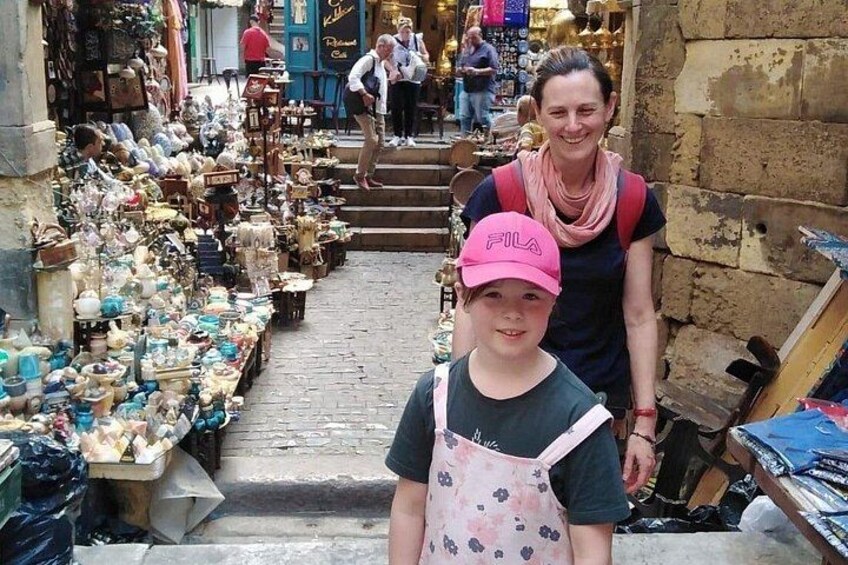 A narrow street with shops on either side, a woman and a child in the foreground, and people walking in the background. 4